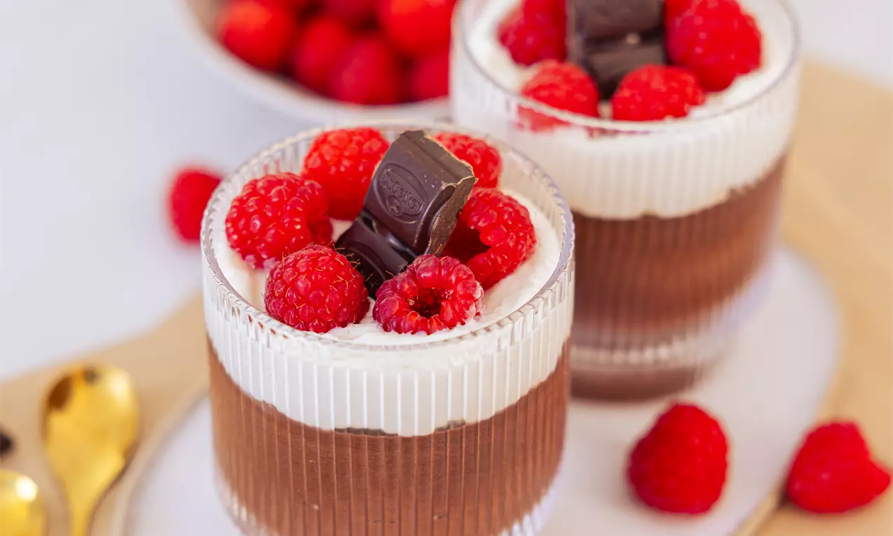 Čokoladni puding v rebrastih steklenih kozarčkih, prekrit s plastjo smetane, svežimi malinami in koščki temne čokolade na beli podlagi