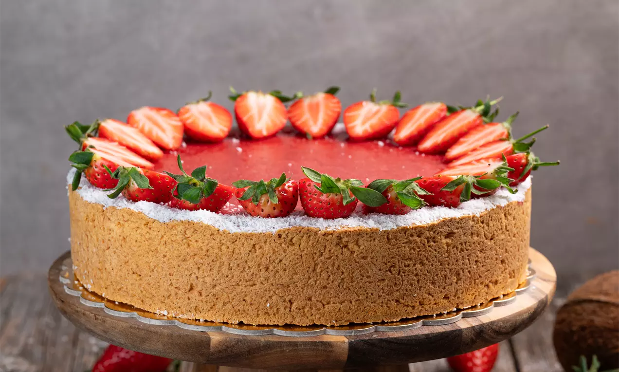 Na lesenem podstavku je krožna kokosova torta z zlatorjavo podlago, gladko rdečo jagodno glazuro in svežimi rezinami jagod po obodu