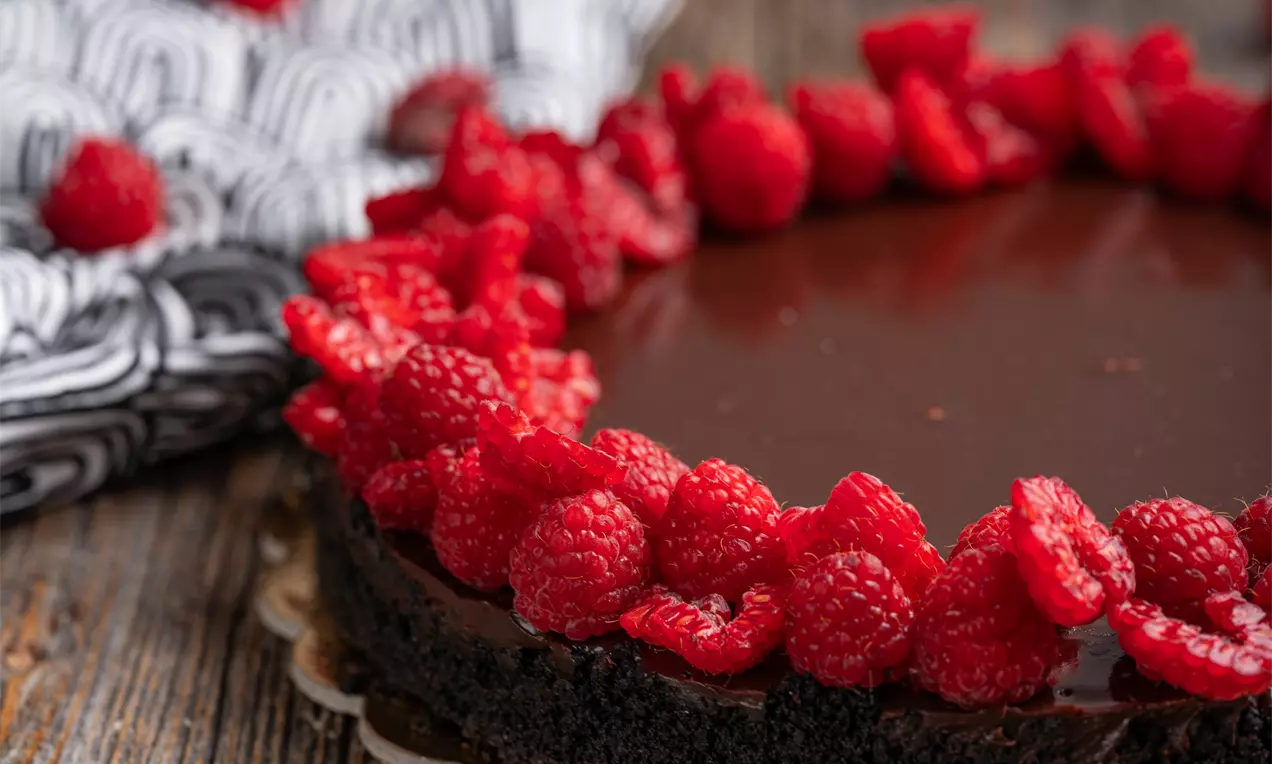Temno rjava čokoladna fudge torta z gladko sijočo površino, obrobljena z živahno rdečimi malinami na rustikalnem lesenem podstavku