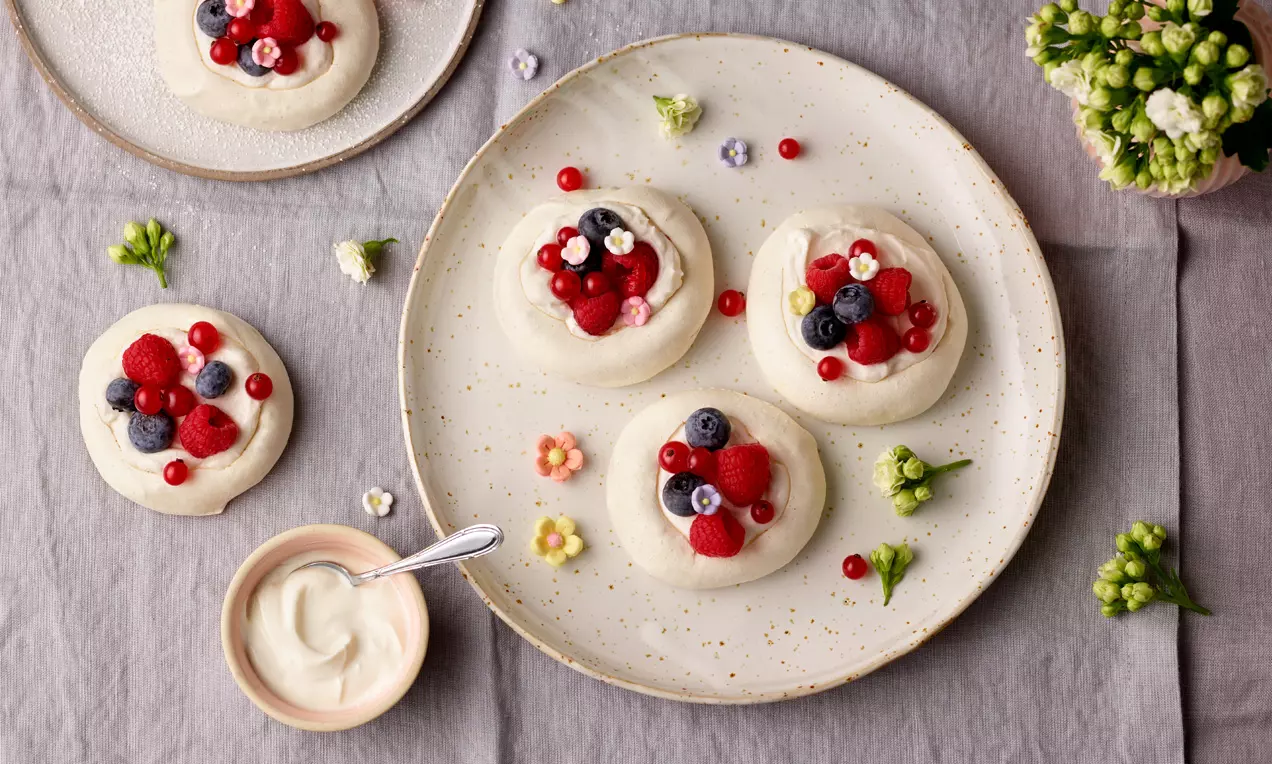 Mini Pavlova svetle kremne barve z gladko površino, obložena z svežimi rdečimi in modrimi jagodami ter postrežena na nežno pikčastem krožniku