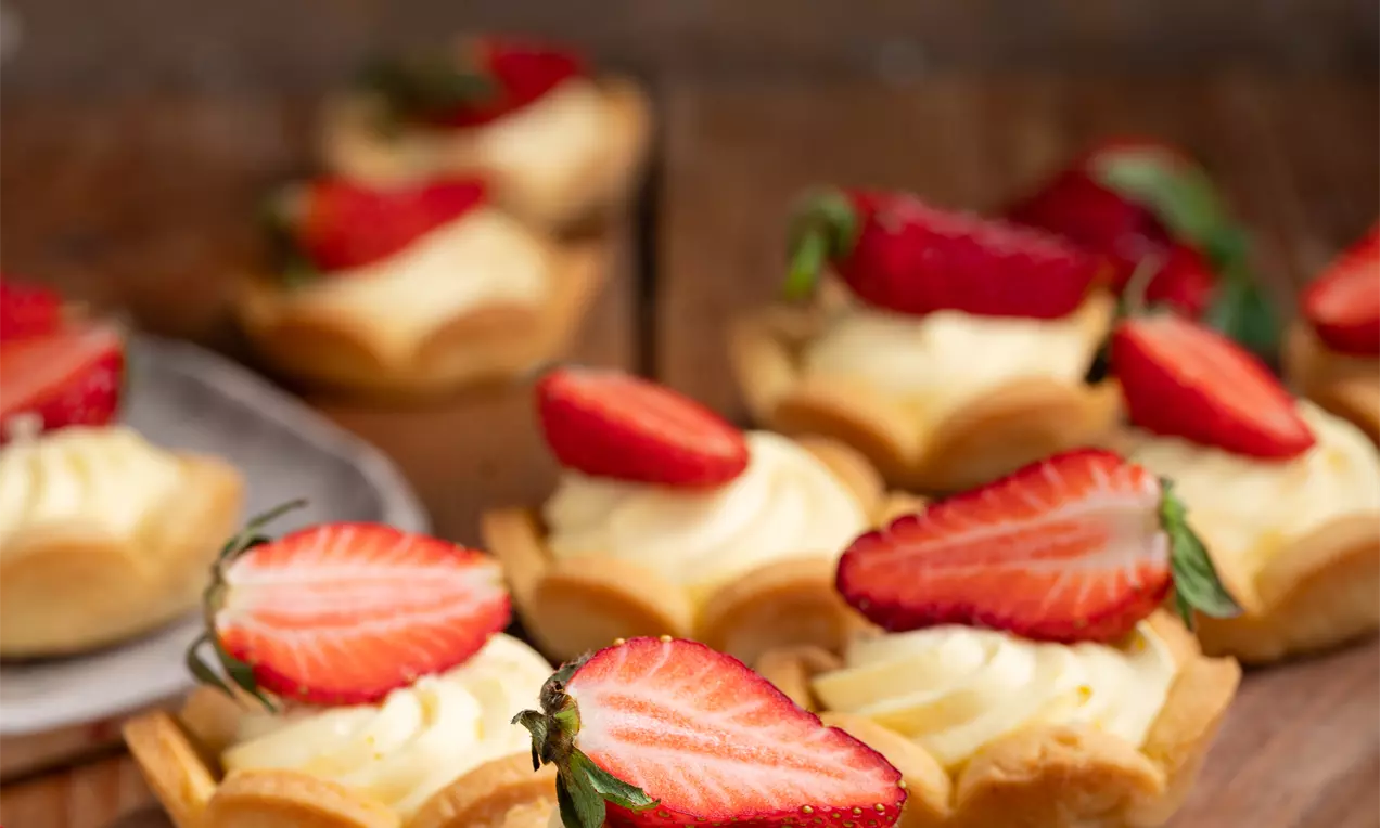 Krhke košarice nežno zapečene barve, napolnjene z gladko vanilijevo kremo in okrašene s svežimi rezinami jagod