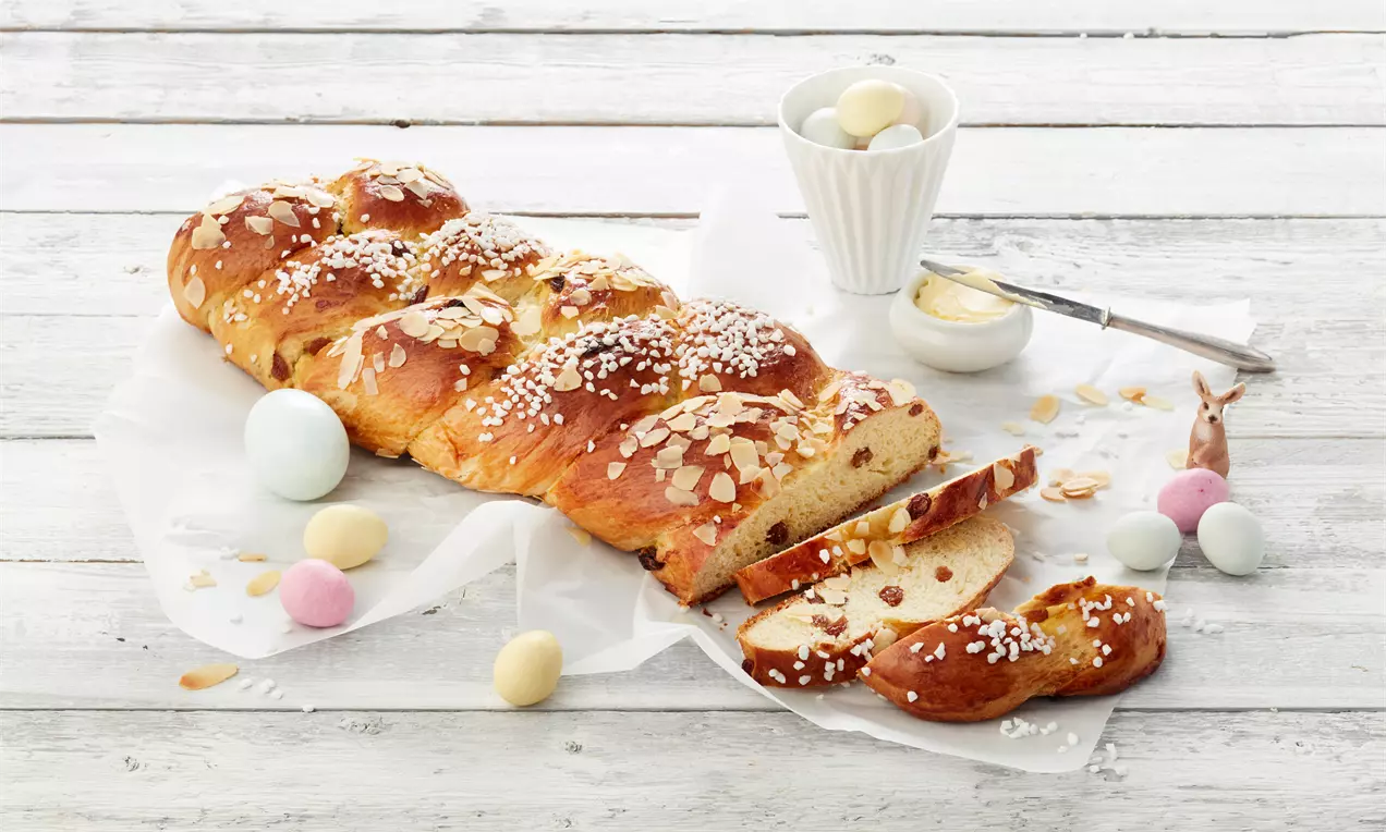 Zlatnosmeđi pleteni kolač s glazurom od krupnog šećera i listićima badema serviran na papiru, uz obojena jaja i bijelu zdjelicu