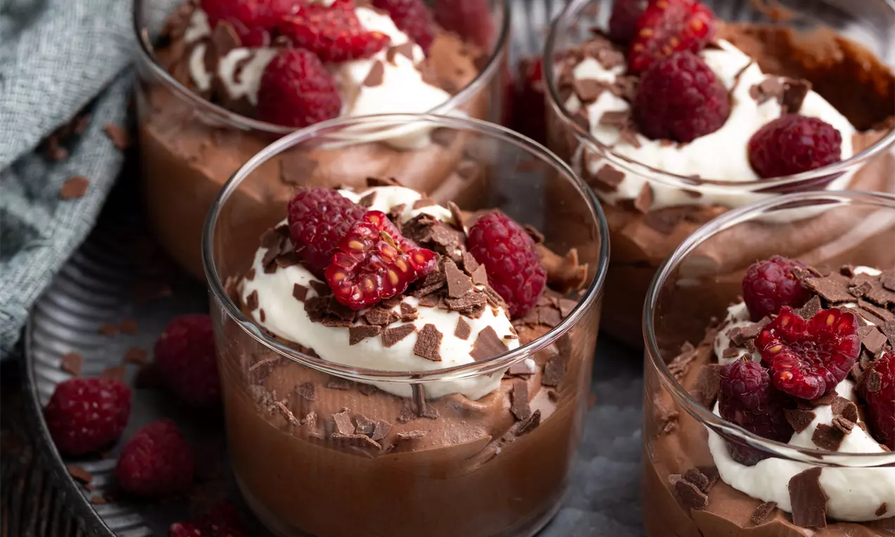 Čokoladni mousse temno rjave barve v prozornih kozarcih, s kremo, malinami in čokoladnimi kosmiči, postrežen na kovinskem pladnju