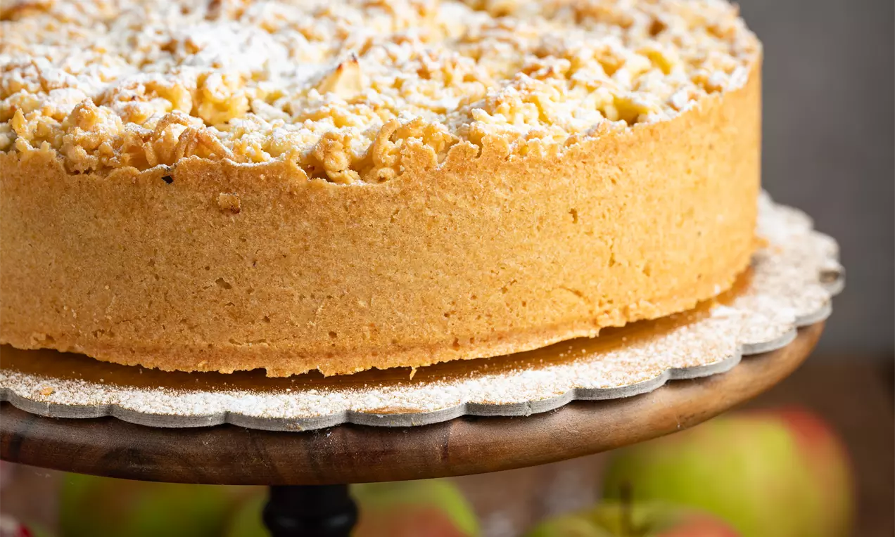 Svetlo rumena torta z zlato rjavim drobljencem na vrhu, posuta s sladkorjem v prahu, postavljena na črnem stojalu, obdana z jabolki