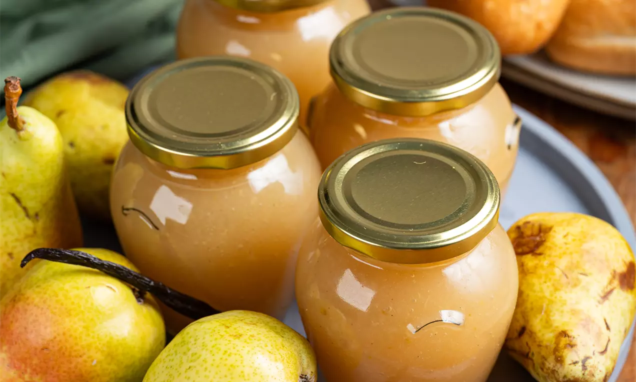 Kozarci z zlato-rjavo hruškovo marmelado z zlato zaporko na modrem krožniku, obkroženi s svežimi hruškami, vaniljevimi stroki in pecivom