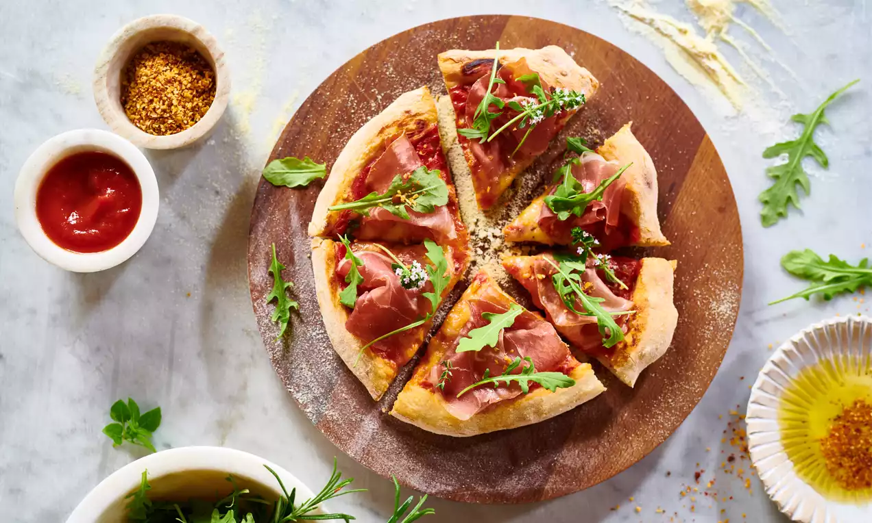 Pica z rjavkasto zlato hrustljavo podlago, obložena s tankimi rezinami pršuta, rdeče paradižnikove omake in svežo rukolo na leseni deski