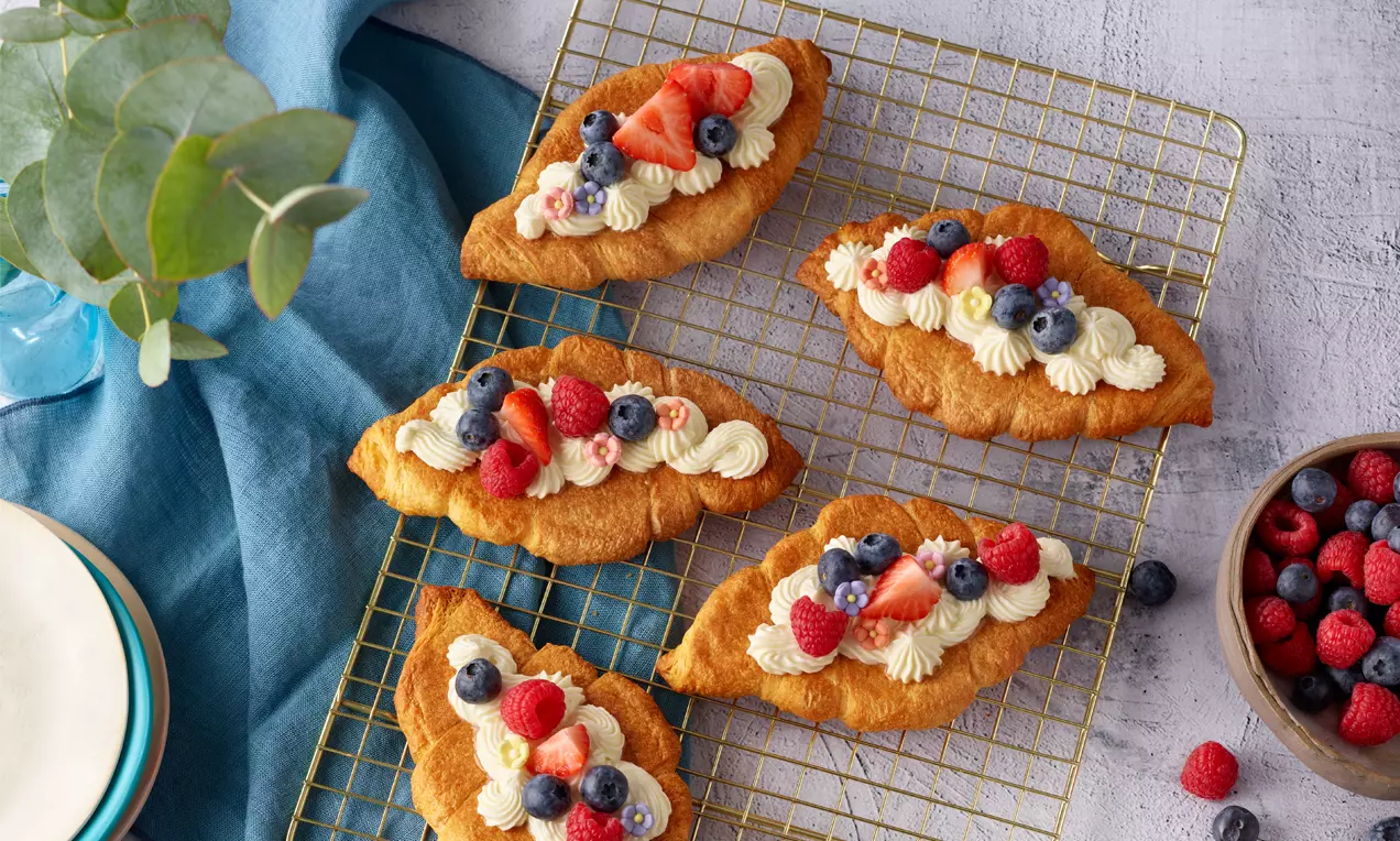 Zlato rjavi roglji v podolgovati obliki, prekriti z nežno smetano in svežimi jagodami, razporejeni na svetlečem kovinskem mrežastem pladnju