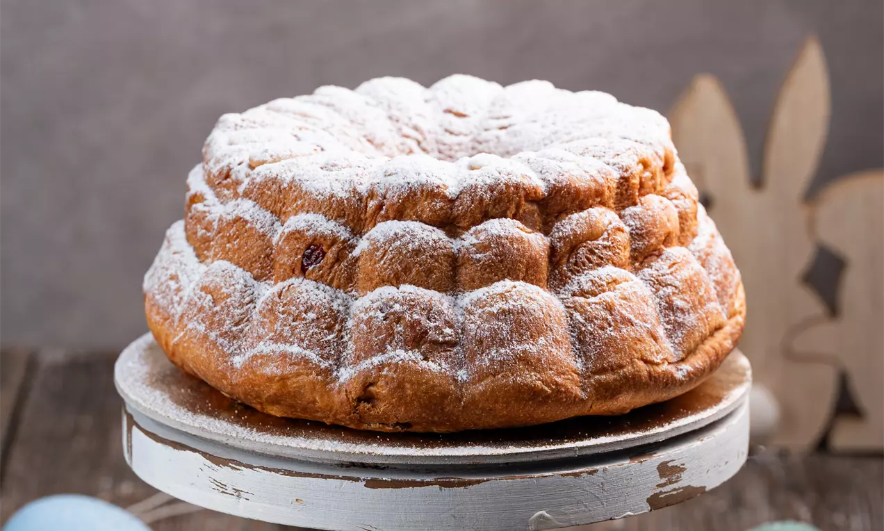Rahlo zlato zapečen kvasen šarkelj z rebrasto teksturo, posut s sladkorjem v prahu, postavljen na lesenem stojalu, obdan z velikonočno dekoracijo