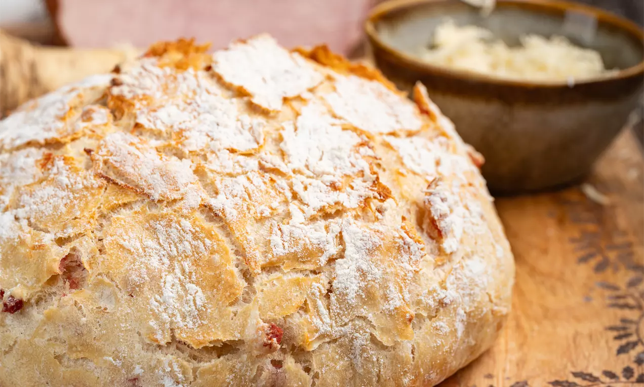 Velikonočni hlebec zlato rjave skorje z nežno razpokano teksturo, posut z moko, na leseni deski ob jajcih in skledi z naribanim hrenom