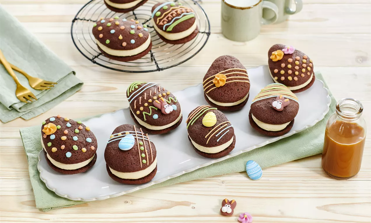 Tamnosmeđi okruglasti kolačići s bijelim punjenjem, ukrašeni šarenim glazurnim detaljima na bijelom tanjiru, okruženi Uskrs dekoracijama