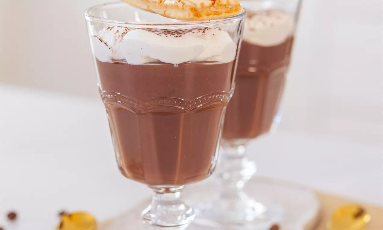 Kremna vroča čokolada temno rjave barve v steklenem kozarcu, okrašena z belo smetano in piškotom, položena na svetlo podlago