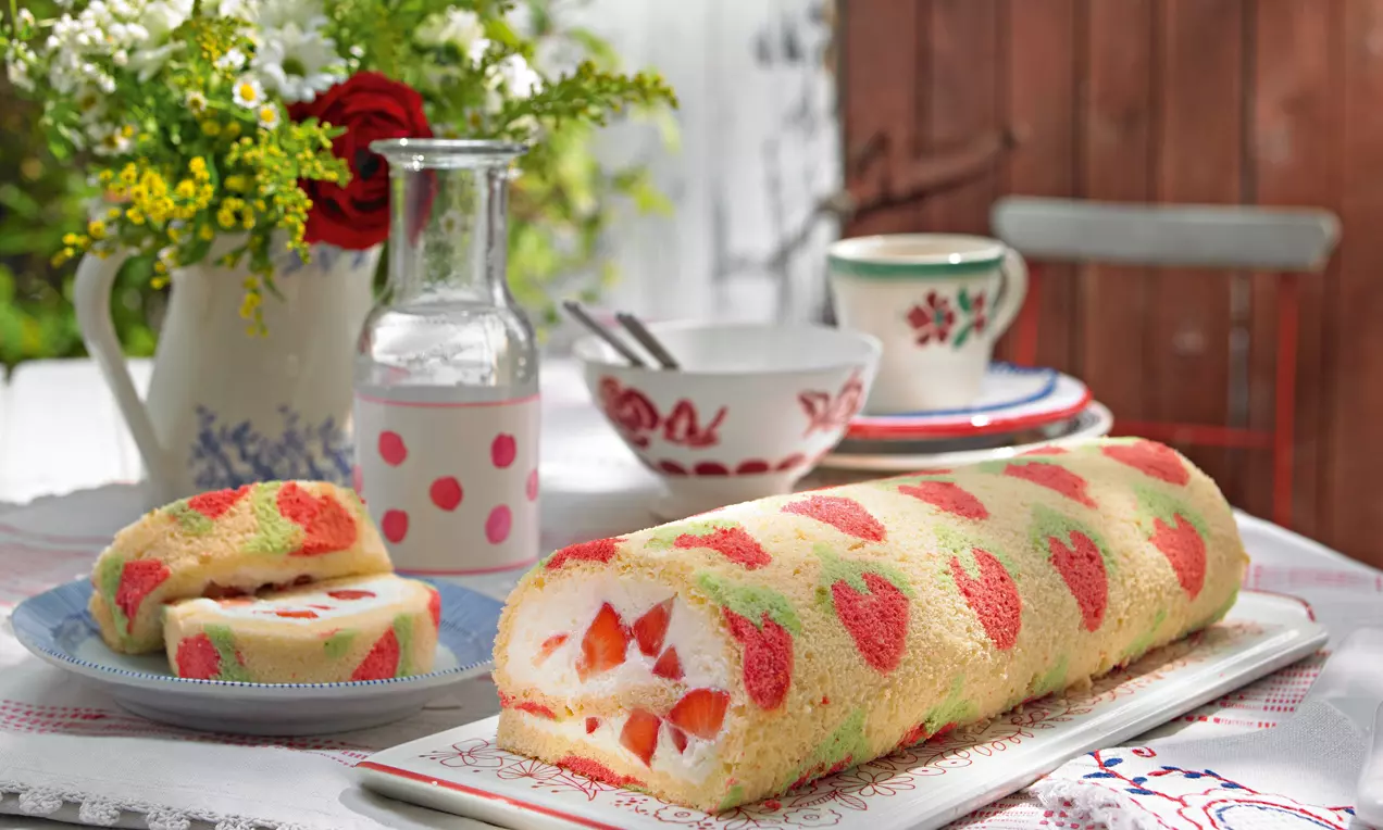 Svetla rumena biskvitna rolada z motivom rdečih jagod, napolnjena s puhasto belo kremo in koščki svežih jagod na krožniku