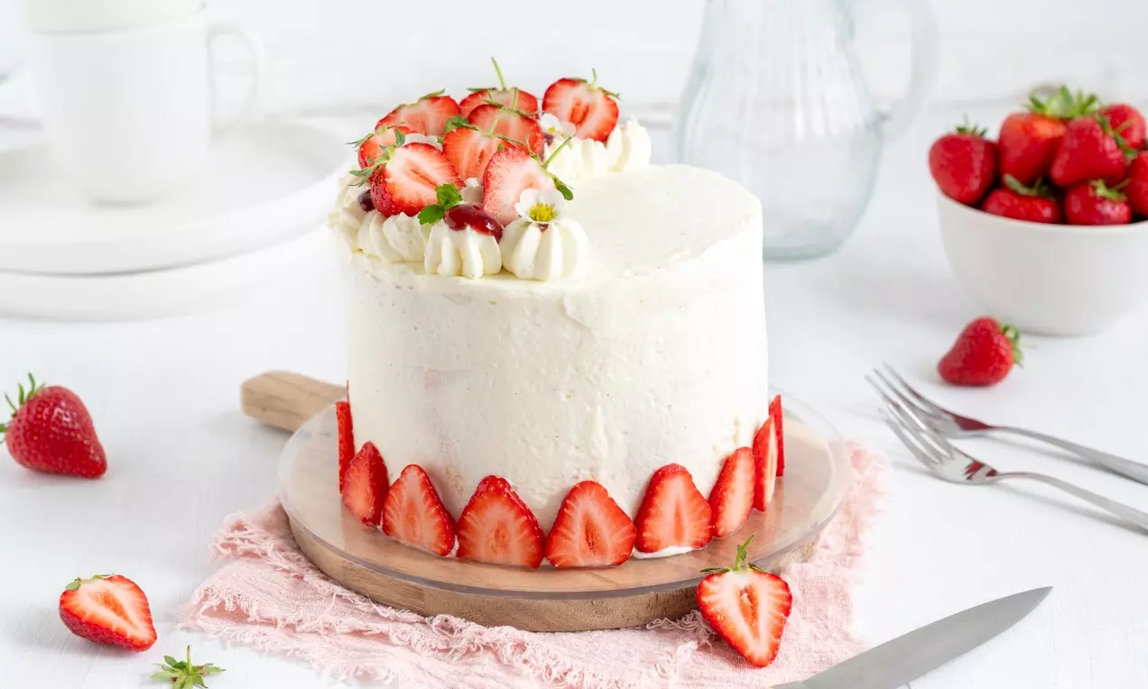 Visoka torta z gladko belo kremo, obrobljena s tankimi rezinami jagod ter okrašena z jagodami in smetanovimi kapicami na leseni podlagi