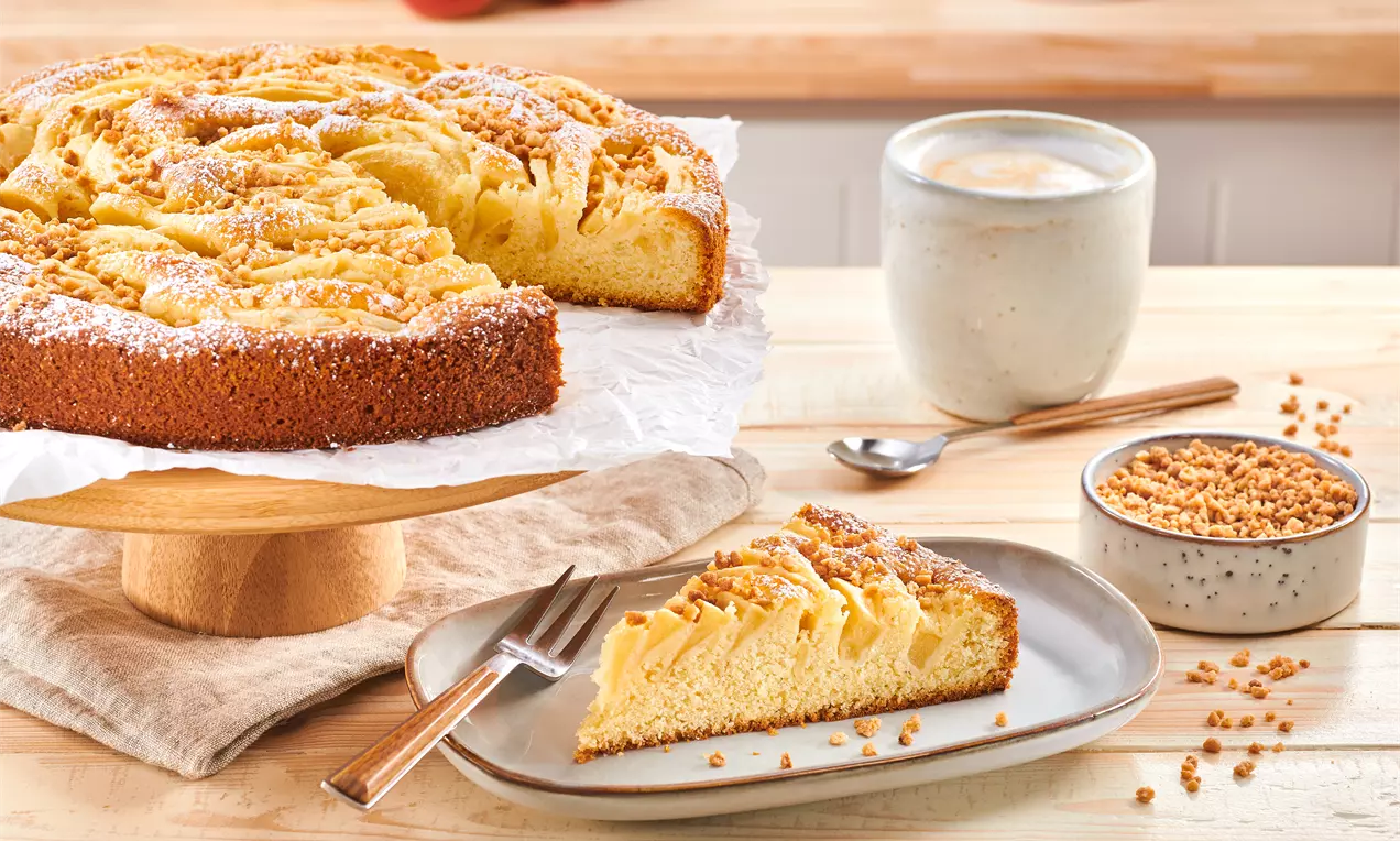 Sočna mandljeva torta z zlato zapečeno skorjico in rezinami jabolk na puhasti podlagi, posuta s sladkorjem v prahu na leseni podlagi.