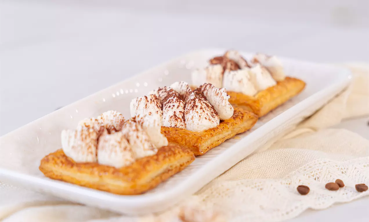 Zlato rjavo listnato testo s kepicami nežne bele kreme, posute s kakavovim prahom, postreženo na belem krožniku z nežno tkanino v ozadju